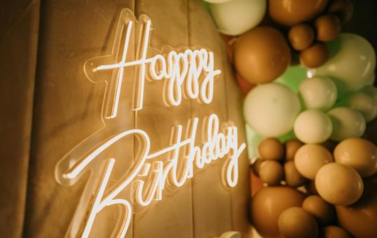 A glowing white neon-style 'Happy Birthday' sign mounted on a warm wood panel, next to a cluster of round balloons in brown, beige, and pale green.