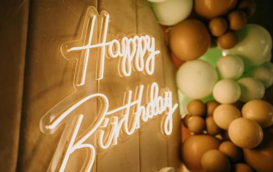A glowing white neon-style 'Happy Birthday' sign mounted on a warm wood panel, next to a cluster of round balloons in brown, beige, and pale green.