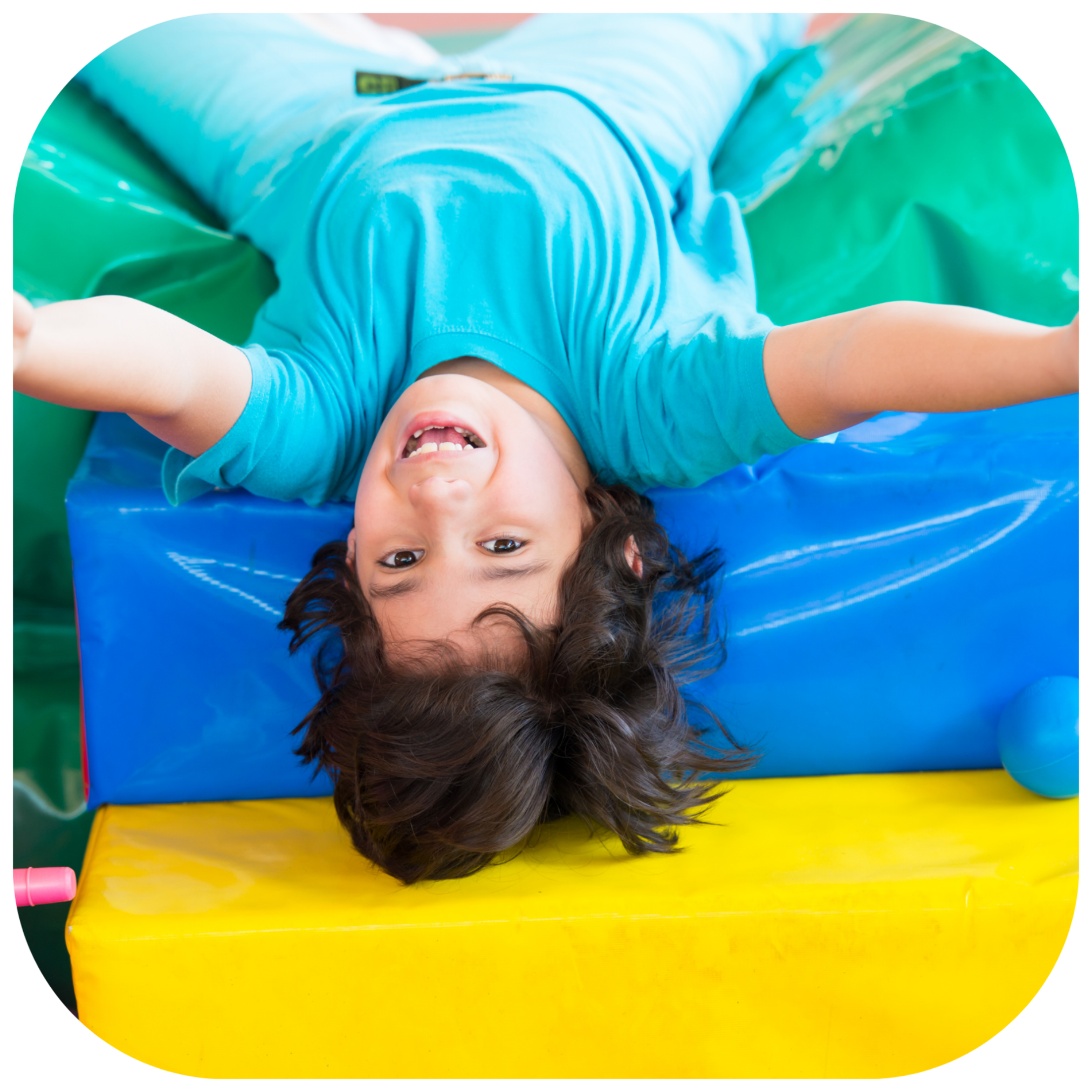 A child wearing a bright turquoise blue t-shirt is lying upside down on colorful foam blocks, including green, blue, and yellow.