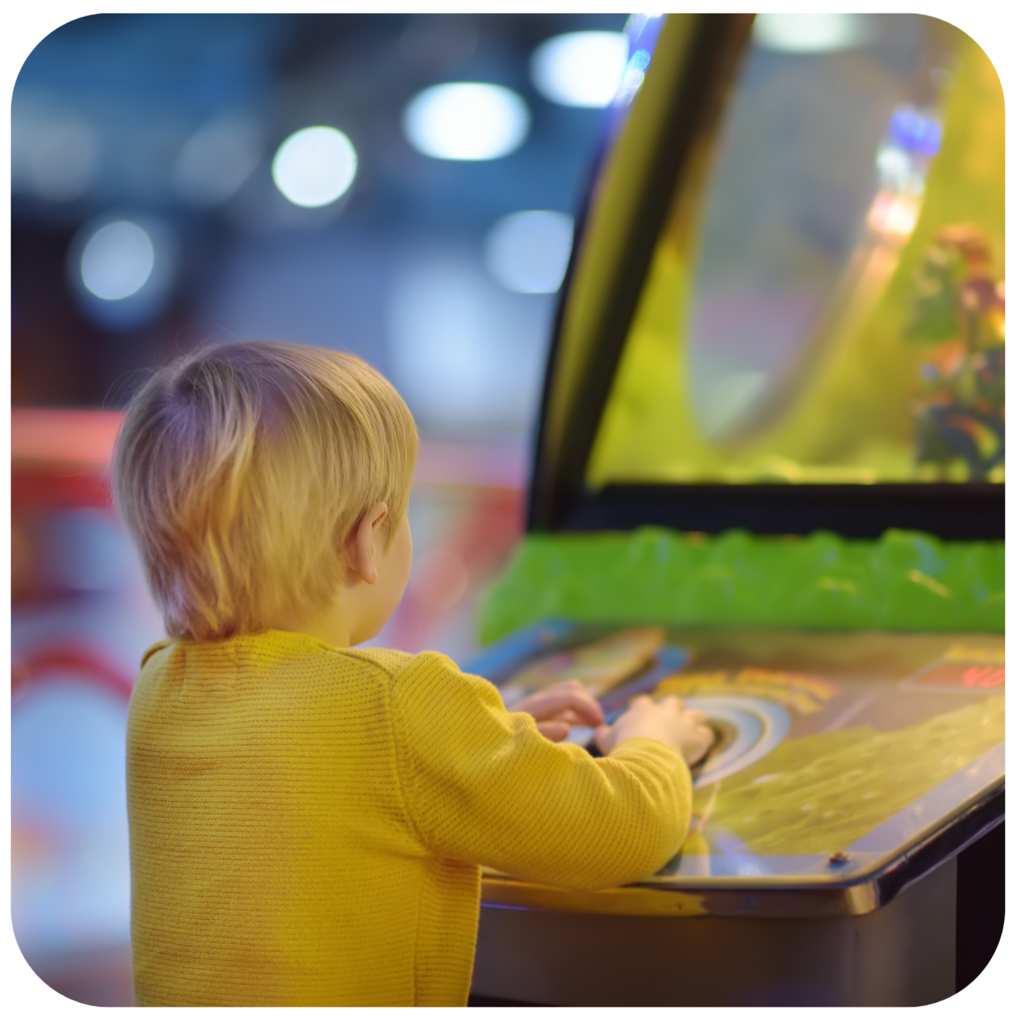 A young child with blonde hair wearing a yellow long-sleeve shirt is seen from behind, playing at an arcade machine with a colorful screen and green trim in a blurred indoor setting.